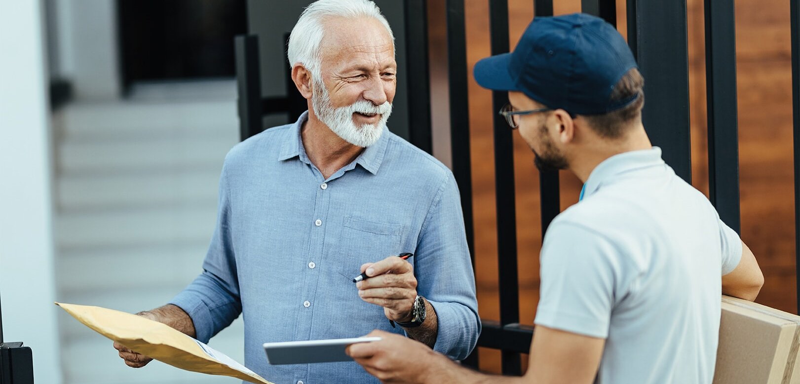 Ein älterer Mann spricht lächelnd mit einem Zusteller, der eine Kiste hält. Der Mann hat ein Umschlag in der Hand und einen Stift.